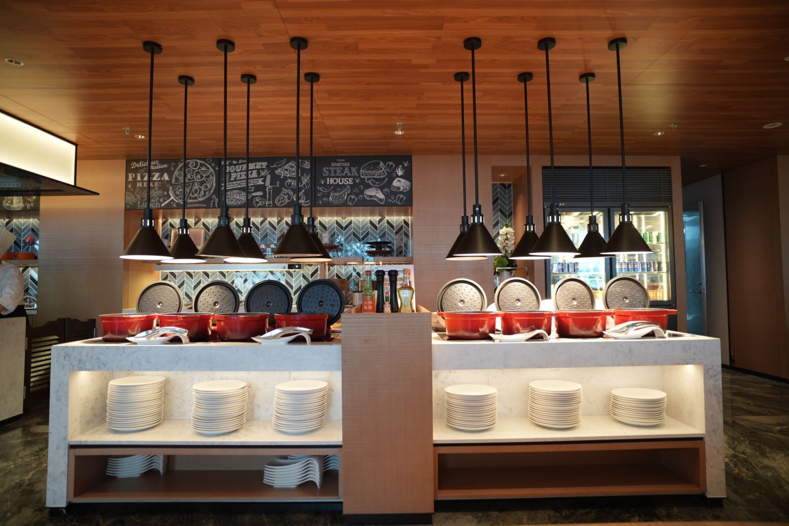 Interior de un restaurante con una barra central que exhibe platos y ollas rojas, iluminación colgante y un panel con ilustraciones de comida.