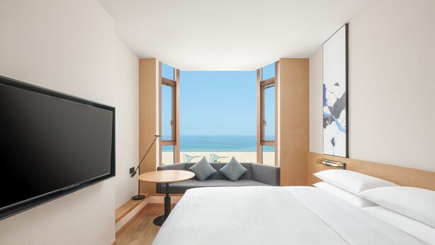 Una camera d'albergo con un letto grande, una TV, un tavolino, un divano con cuscini e vista mare attraverso finestre aperte.