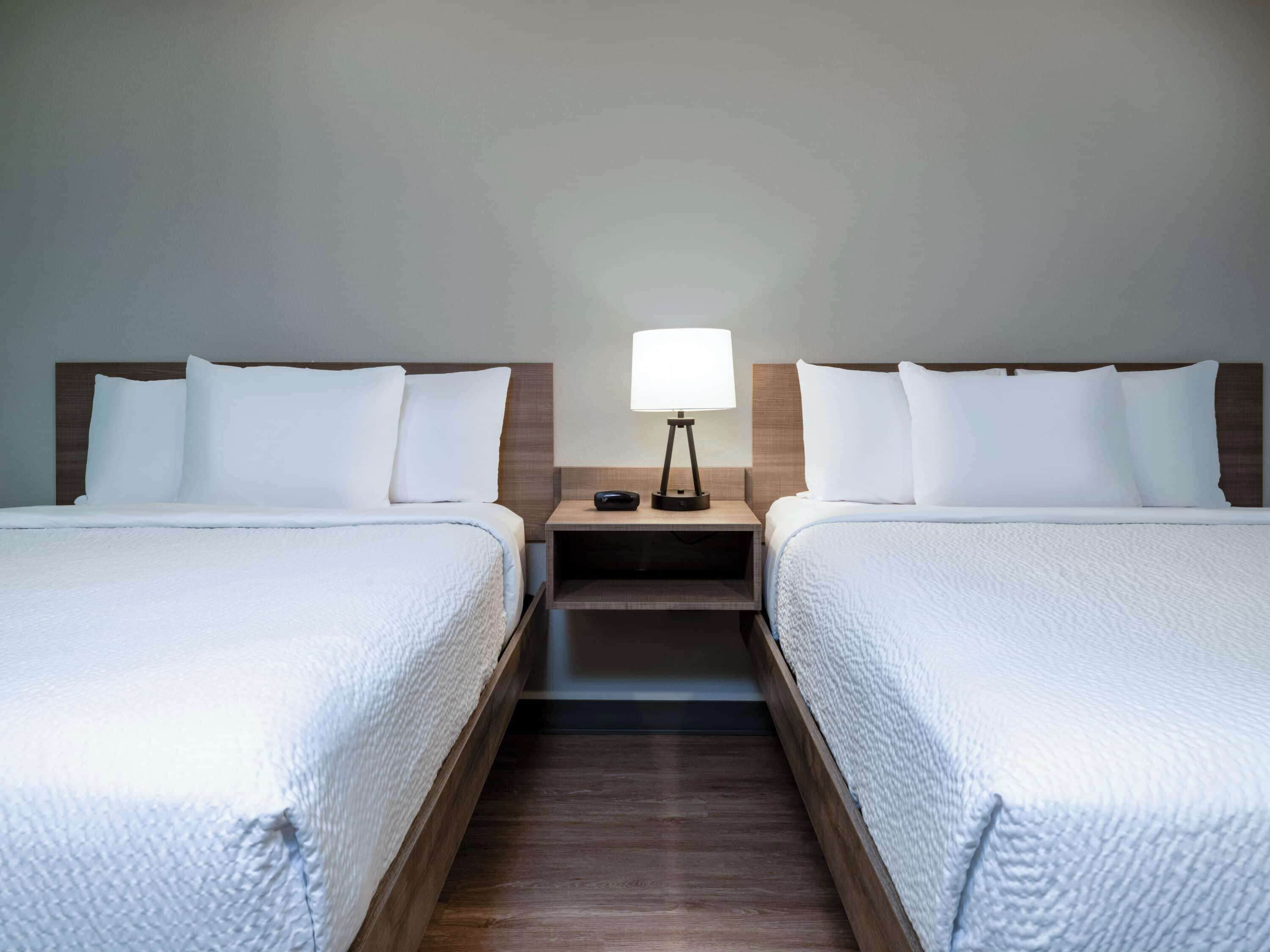 Hotellihuone, jossa on kaksi sänkyä, yöpöytä lampun kanssa ja puulattia.