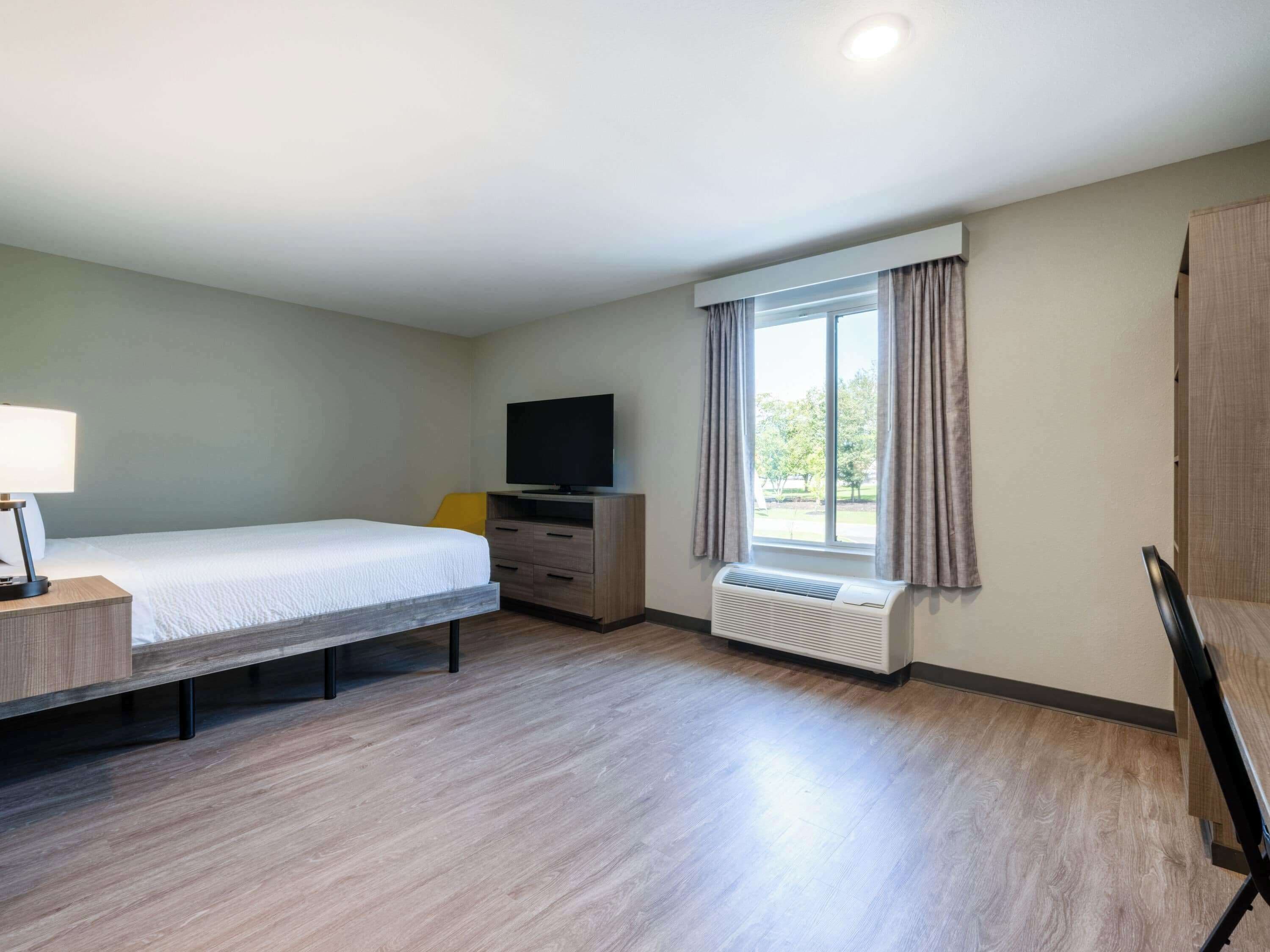 A hotel room with a bed, a TV, a desk with a lamp, a window with curtains, and a radiator.