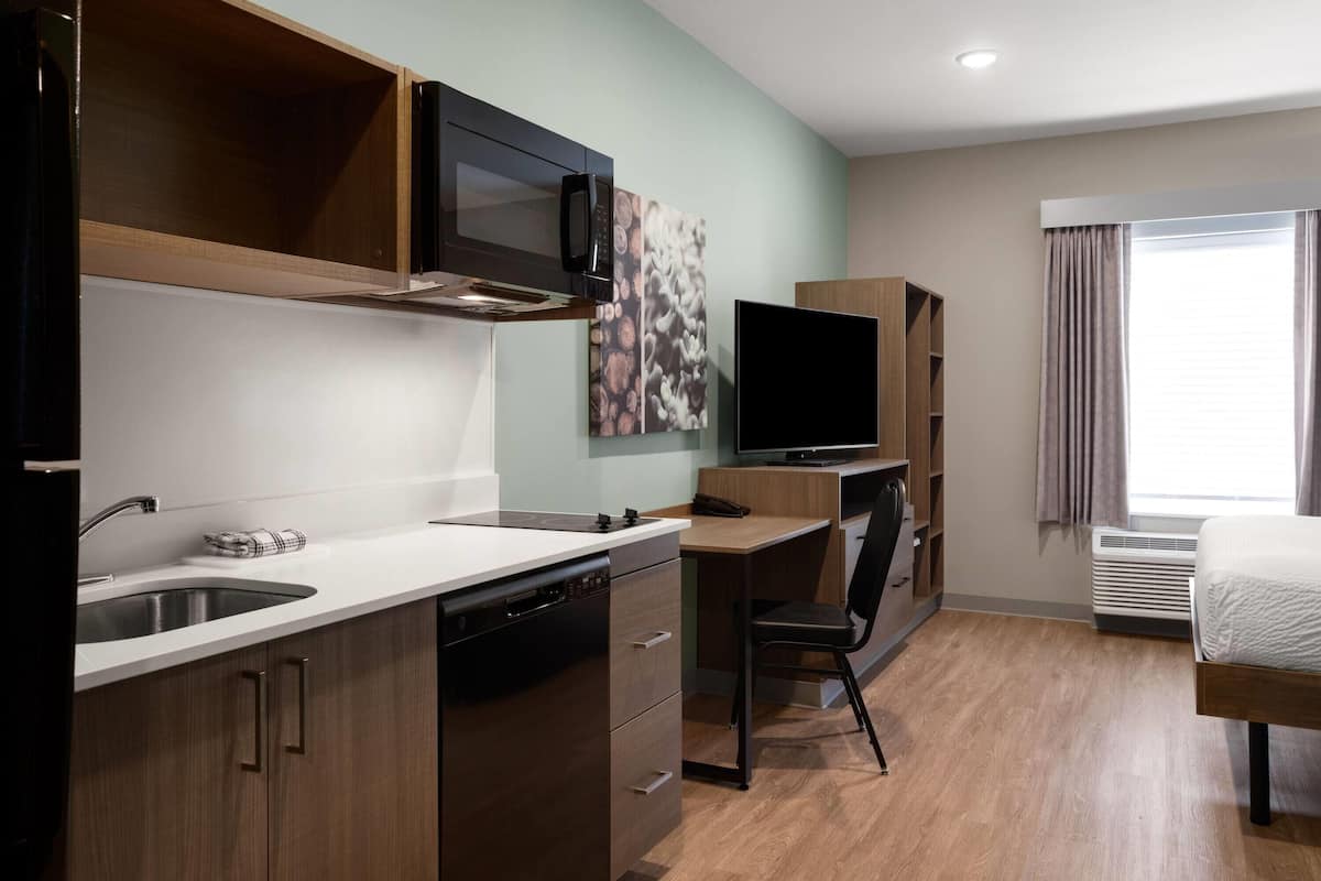 A compact hotel room with a kitchenette, a bed, a desk, and a TV.