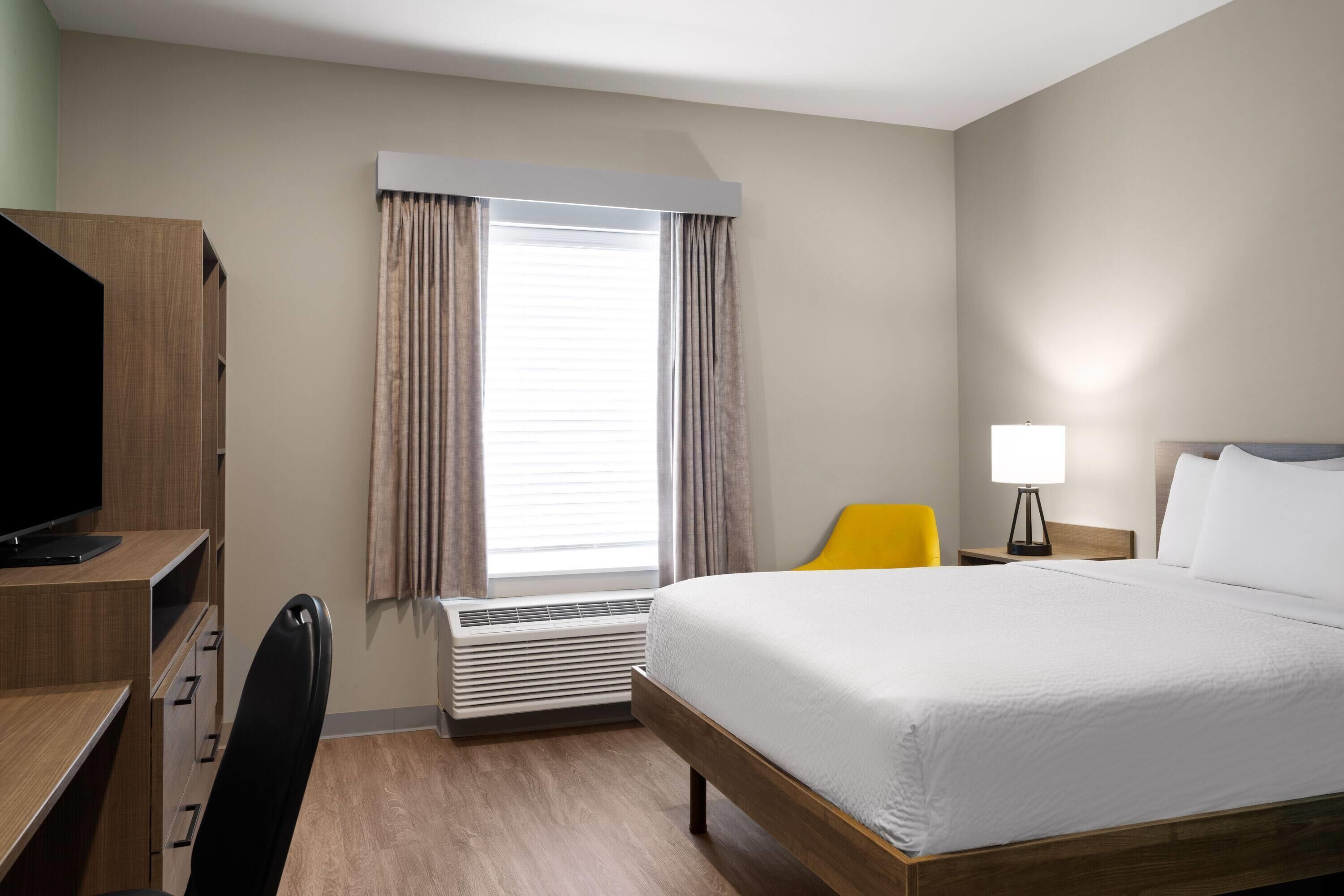 A hotel room with a bed, a desk with a computer, a chair, a lamp, and a window with curtains.