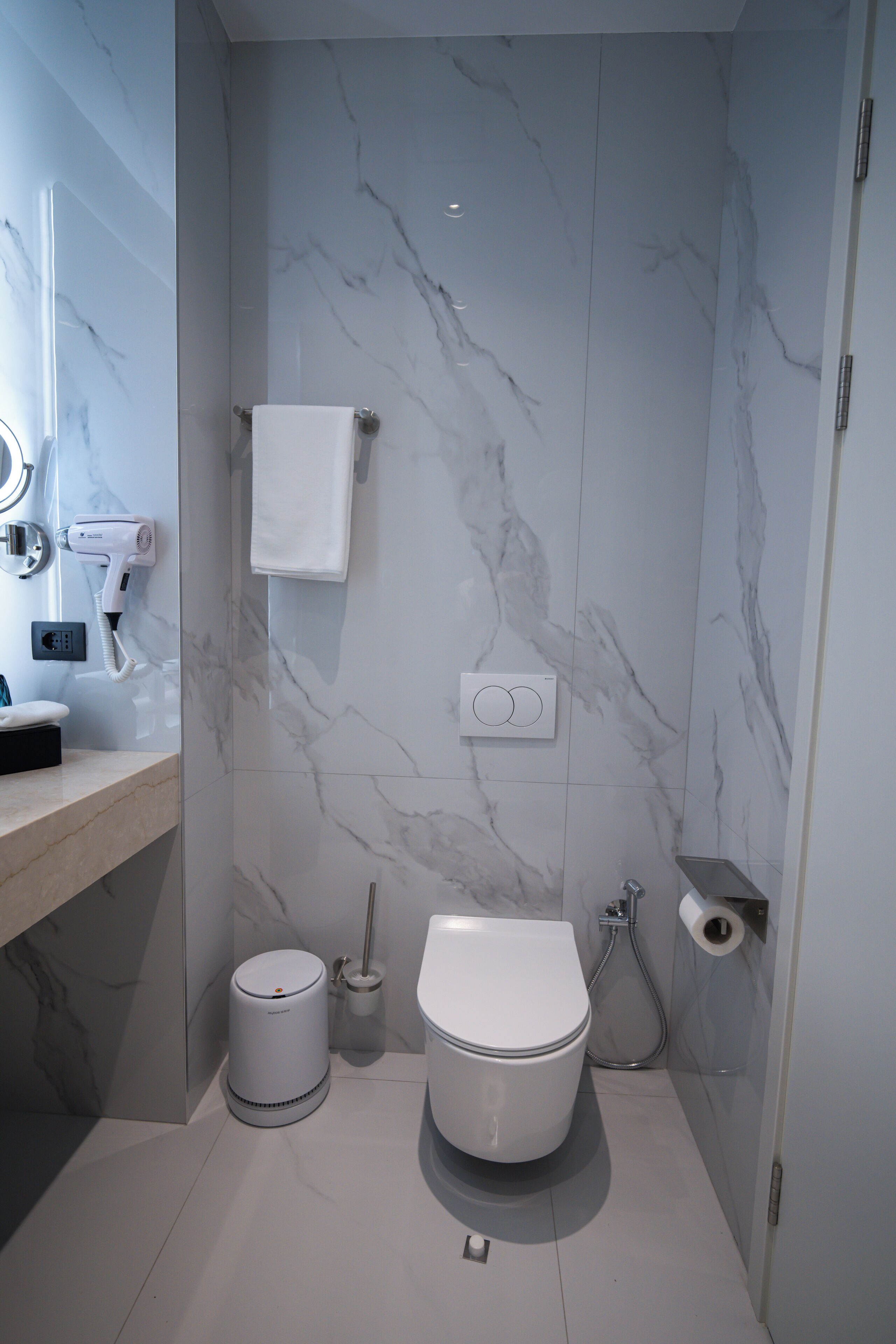 A modern bathroom with a marble wall, a toilet, a bidet, a hairdryer, and a trash bin.