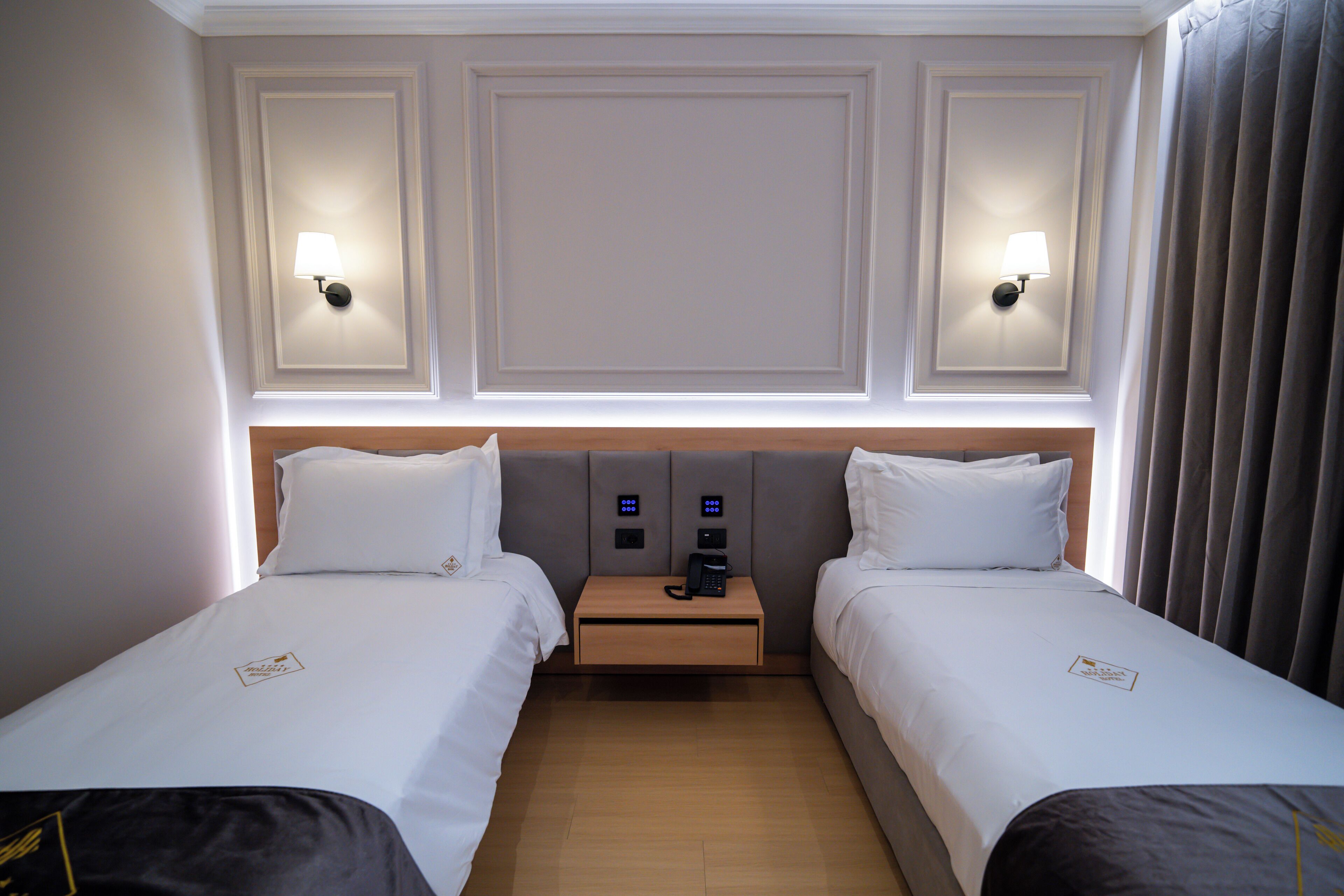 A hotel room with two beds, a nightstand with a phone, and wall-mounted lamps.