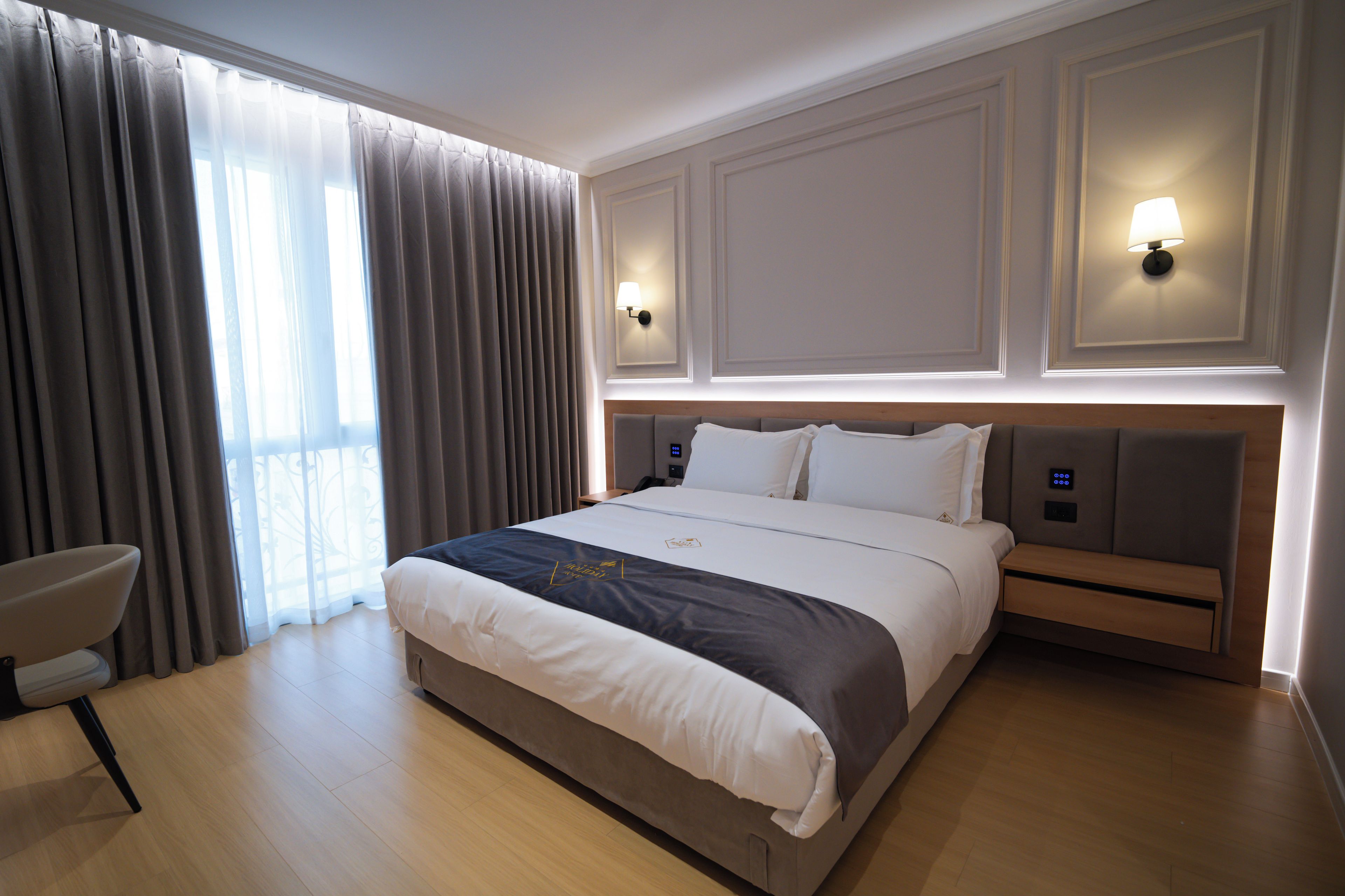Habitación de hotel con una cama grande, dos lámparas de noche, una silla y vistas al exterior a través de cortinas traslúcidas.