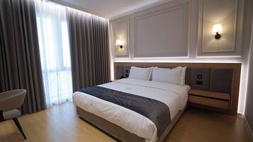 Habitación de hotel con una cama grande, dos lámparas de noche, una silla y vistas al exterior a través de cortinas traslúcidas.