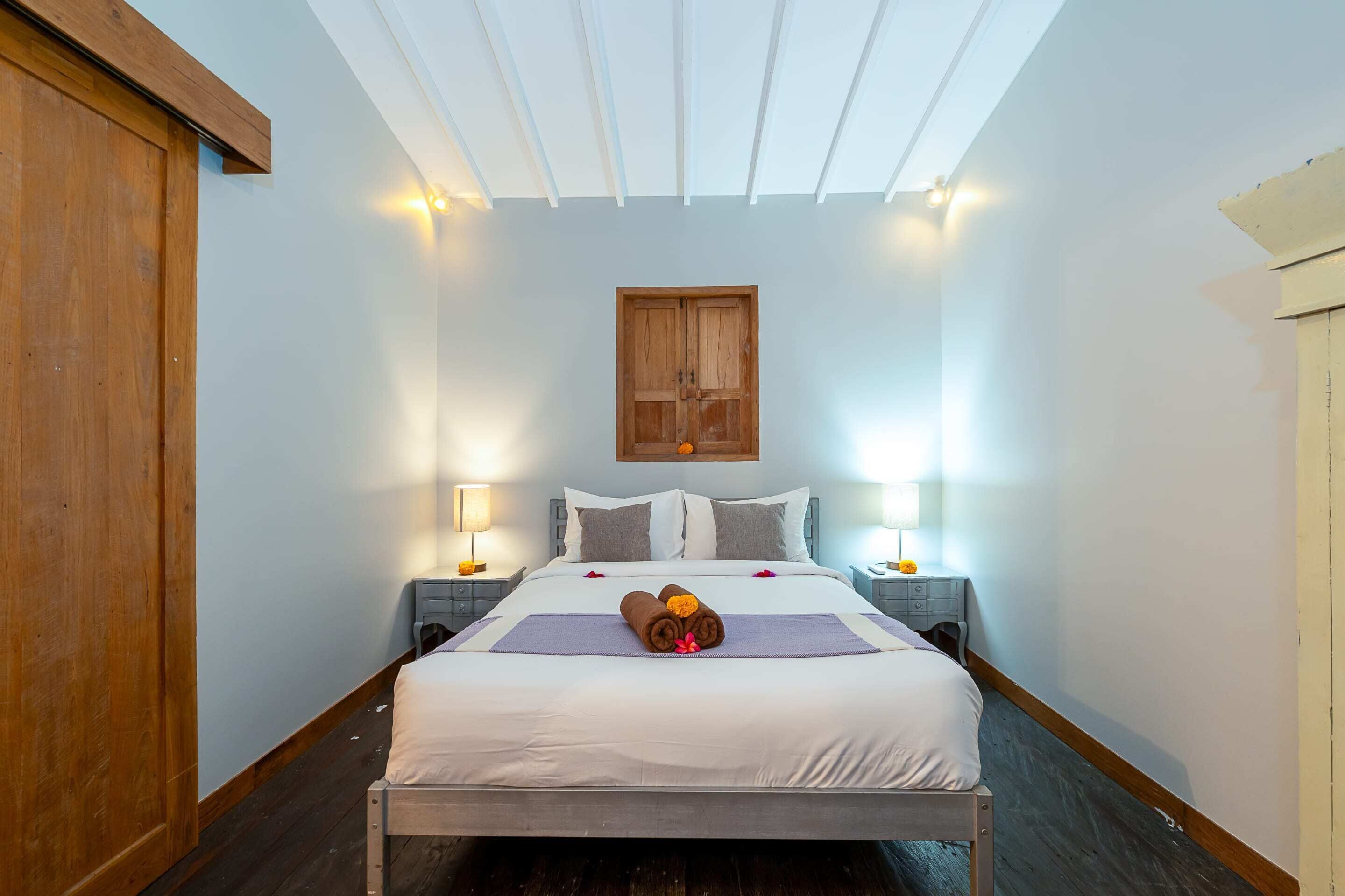 A neatly made bed with a white duvet, two bedside tables with lamps, a wooden wardrobe, and a wooden door.