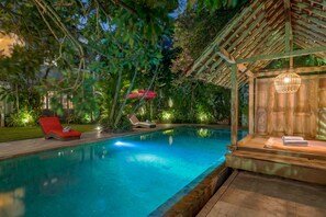A pool area with a wooden deck, lounge chairs, and a pergola with string lights. - Villa Tap (Canggu)
