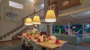 Un coin repas avec une table en bois dressée pour quatre personnes, entourée de chaises blanches. Des lampes sont suspendues au-dessus de la table, et un escalier est visible au fond.