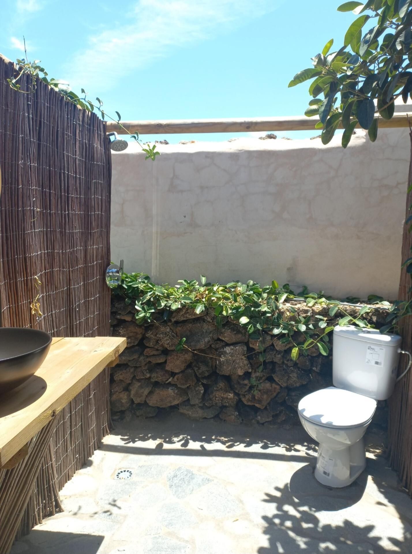 A small outdoor bathroom with a toilet, a stone wall, and a bamboo fence.