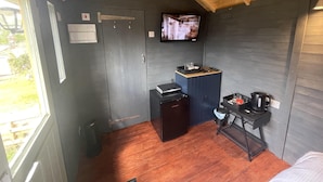 A compact room with a wooden ceiling, a television mounted on the wall, a small kitchenette with a coffee maker, and a window providing a view of the outdoors. - Enchanted Cabin Retreat in Sibford Gower (Banbury)