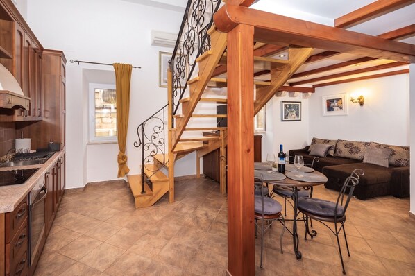 A kitchen with wooden cabinets, a dining area with a table and chairs, a staircase with a wooden railing, and a living area with a sofa and cushions. - Amoret Apartments (Dubrovnik)