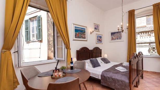 Una habitación con cama, una mesa de comedor con sillas, un ventanal con cortinas y una obra de arte colgada en la pared.
