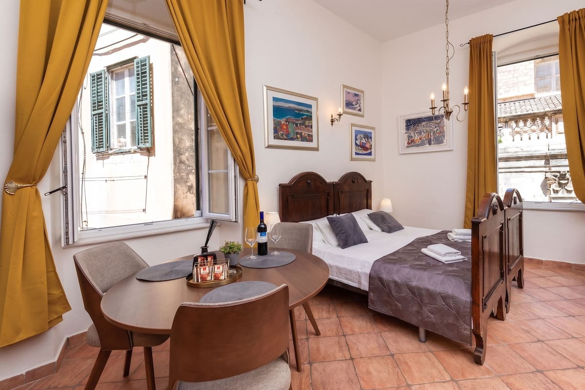 Una habitació amb un llit, taula de menjador amb cadires, finestra amb cortines i obra d'art penjada a la paret.