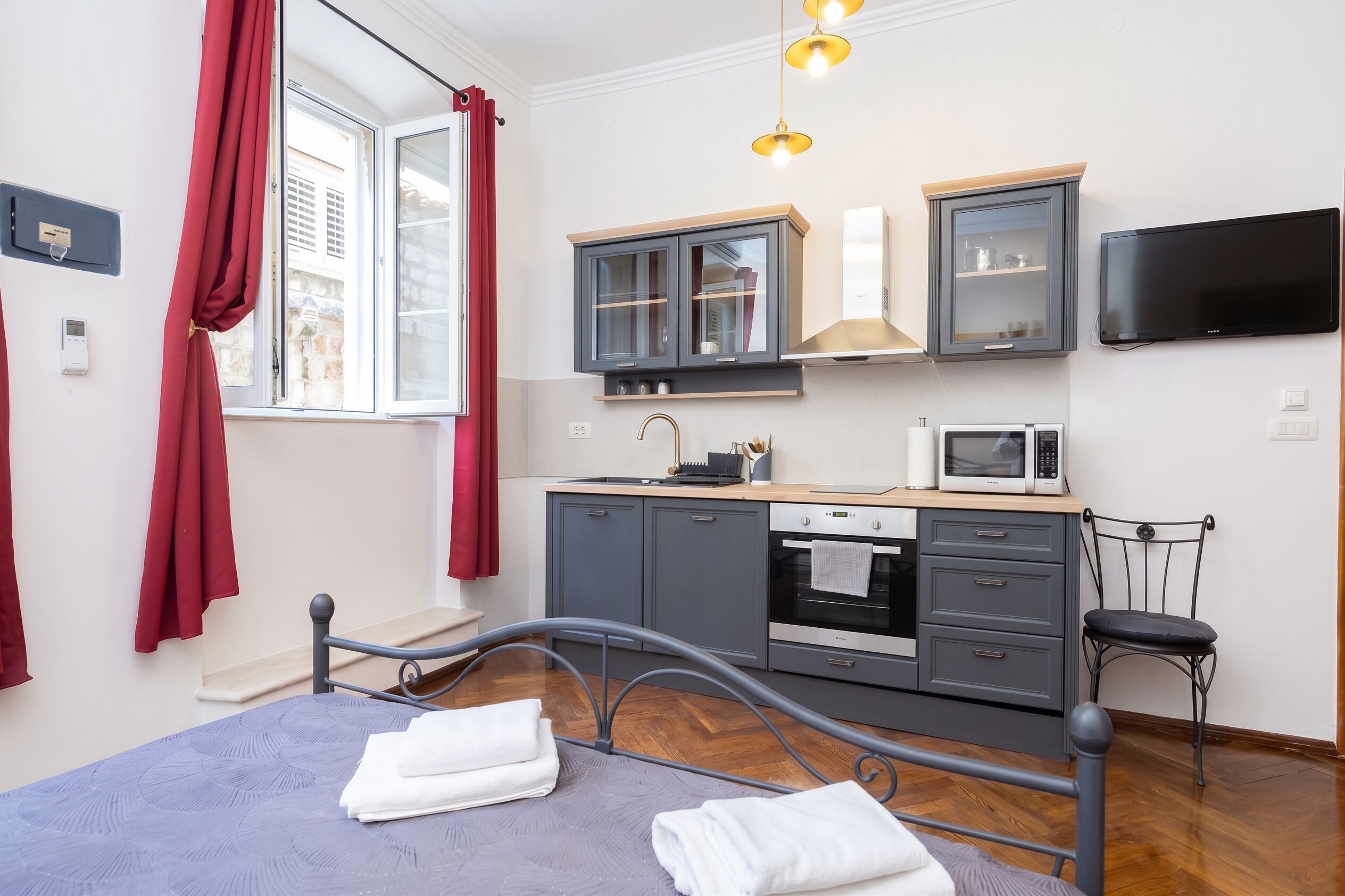 Habitación compacta con cama, mesita de noche, ventana con cortinas rojas, kitchenette con microondas, horno y armarios, y un televisor montado en la pared.
