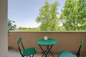 A balcony with two green chairs and a small round table, a white candle, and a view of trees and buildings.