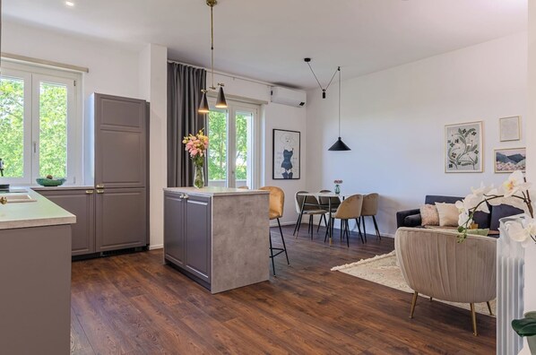 A modern kitchen with a central island, dining area with chairs, and a living room with a sofa and decorative items.