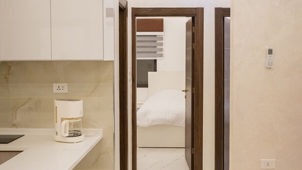 Una cocina moderna con cafetera, una encimera y un pasillo que conduce a un dormitorio con cama y mesita de noche.