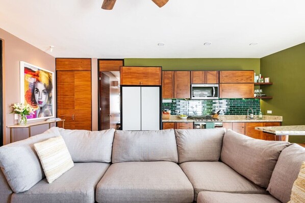 A modern living room with a sectional sofa, a dining table, and a kitchen with wooden cabinets and a green backsplash.