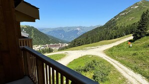 Un balcone in legno affaccia su un paesaggio montuoso con un sentiero sterrato e un edificio in lontananza.