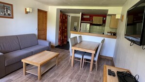 Un ambiente compatto con divano, tavolino in legno, tavolo da pranzo con sedie, angolo cottura con mobili e televisore montato a parete.