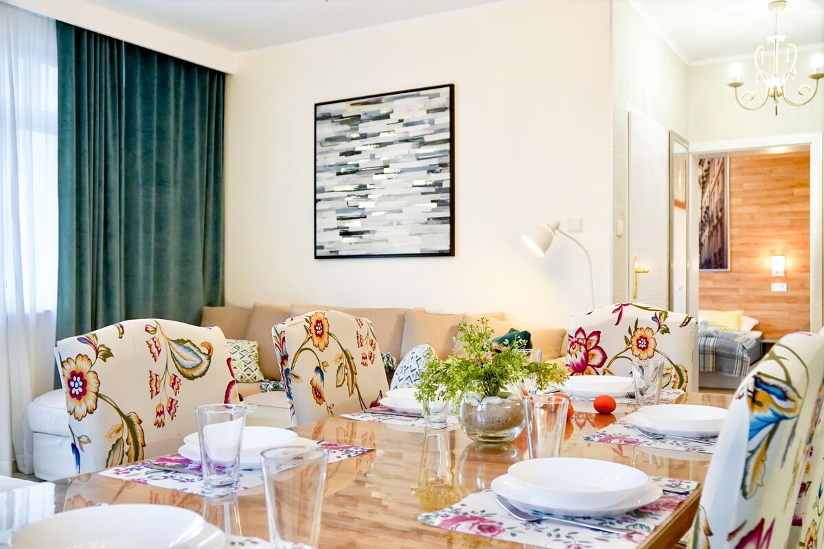 Un coin repas avec une table en bois dressée pour quatre personnes, entourée de chaises à motifs floraux. Un grand tableau abstrait orne le mur, et un lustre est suspendu au-dessus de la table.
