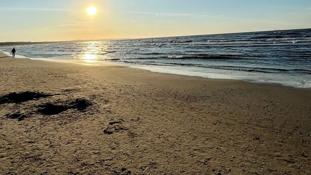 Una playa con el sol poniéndose sobre el océano, orilla arenosa e huellas en la arena.