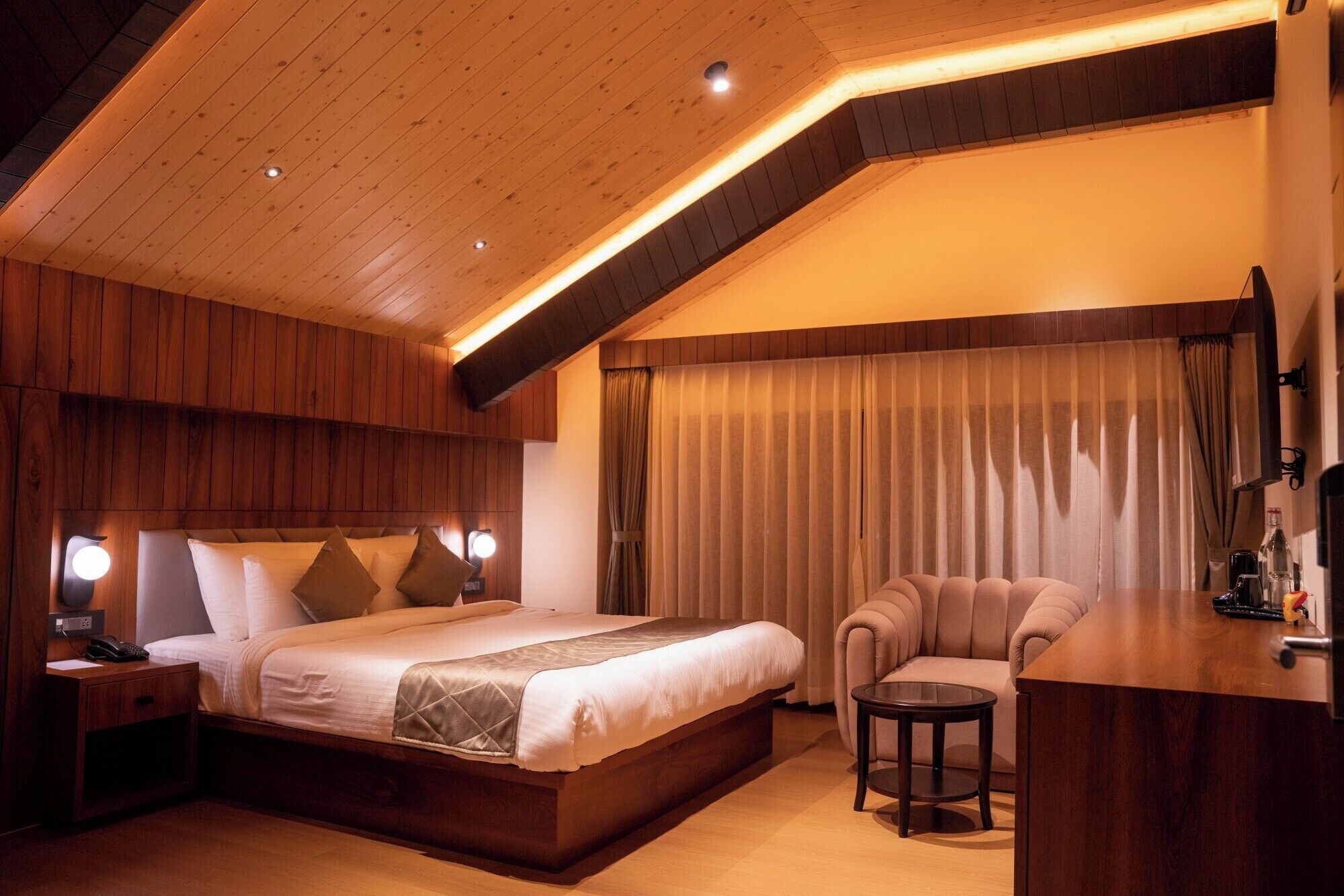 Habitación de hotel con techo de madera, una cama grande con colcha, una silla, una mesita y un escritorio con lámpara.