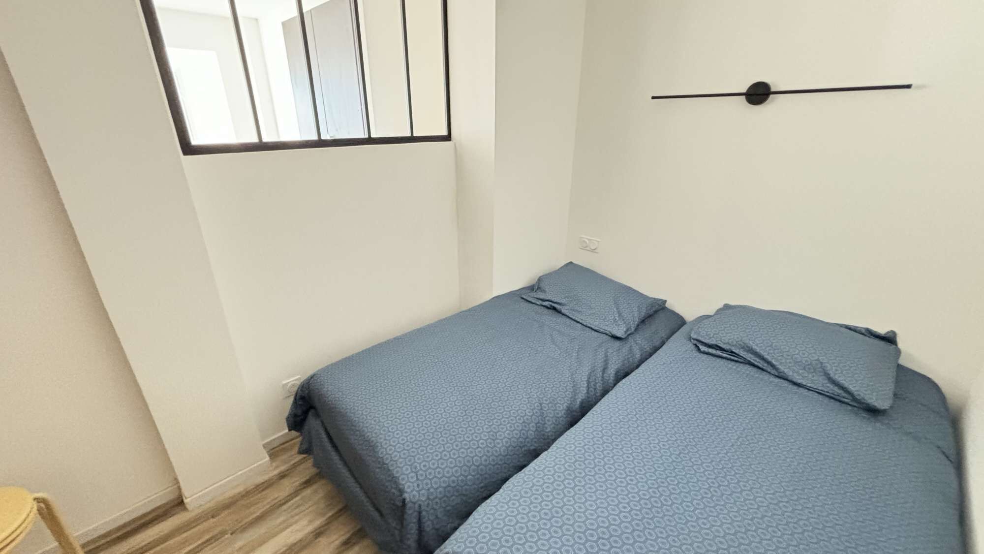 Ein schlichter, kleiner Raum mit zwei Einzelbetten, einem Fenster mit Gitter und einer Wandleuchte.