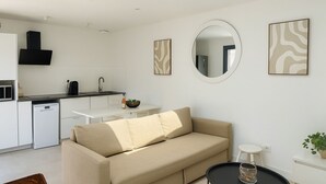 A modern, minimalist living space with a sofa, a dining table, a kitchen area, and a round mirror. - House Garden Pool – The Cherry Tree (Venelles)