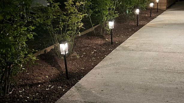 A pathway lined with small, evenly spaced, illuminated lantern-style lights, leading towards a wooden structure with a sloped roof. - House Garden Pool – The Cherry Tree (Venelles)