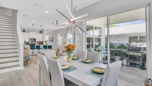 A modern kitchen with white cabinets, a dining area with a white table set for four, and a large glass sliding door leading to an outdoor patio. - Arlington Ocean Oasis 5br 5ba Htd Saltwater Pool (Sarasota)