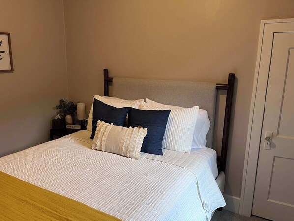 A neatly made bed with a white and blue quilt, a wooden headboard, and a bedside table with a lamp and a plant. - Revitalized Mid-Century Modern Gem (Sioux Falls)