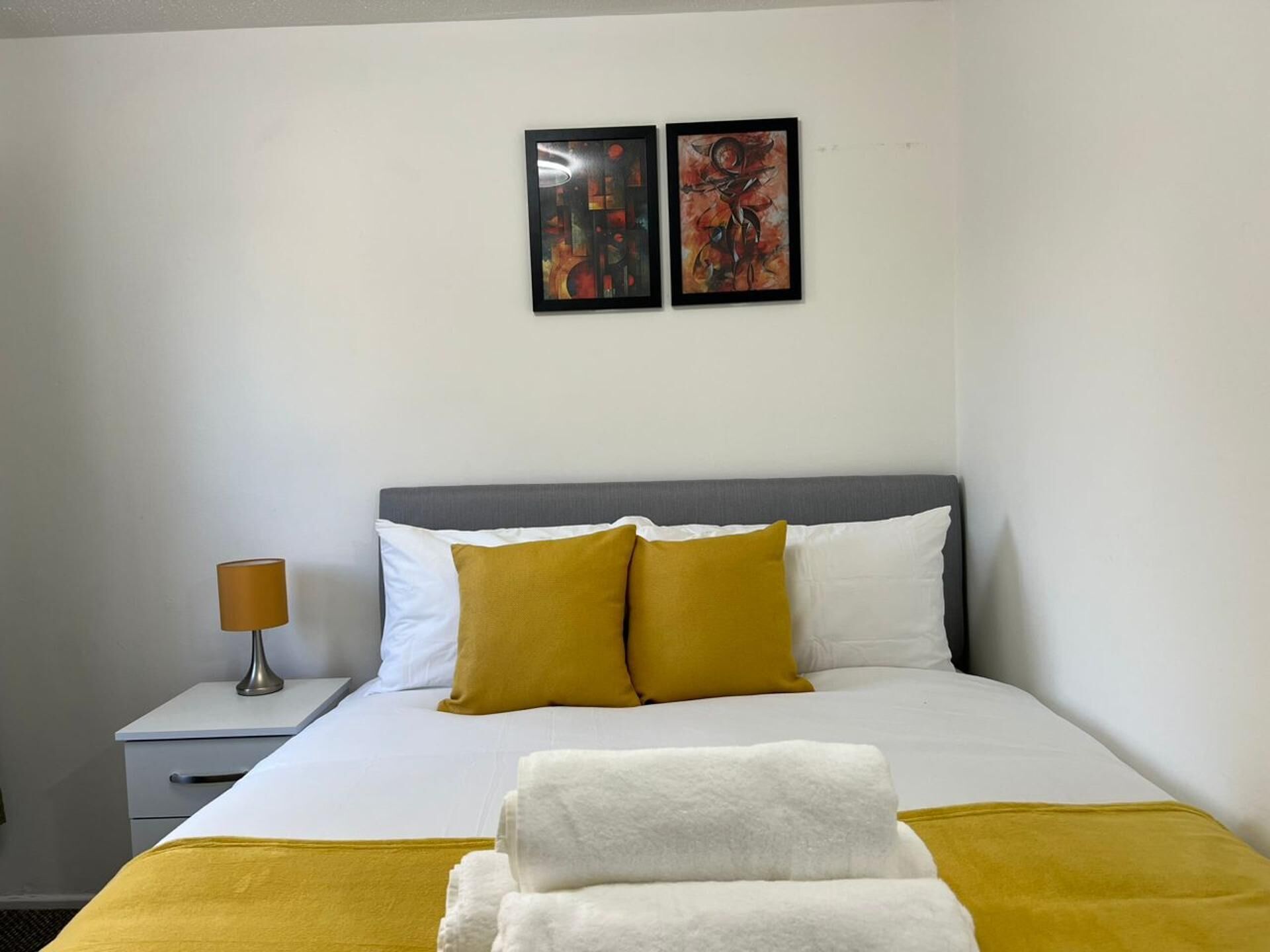 Ein Hotelzimmer mit einem Bett, zwei Kissen, einem Nachttisch mit Lampe und zwei gerahmten Kunstwerken an der Wand.
