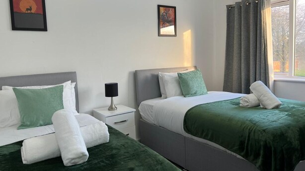 A hotel room with two beds, a nightstand, a lamp, and two framed pictures on the wall. - Birmingham Luxury House - Modern Retreat (Birmingham)