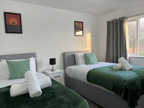 A hotel room with two beds, a nightstand, a lamp, and two framed pictures on the wall. - Birmingham Luxury House - Modern Retreat (Birmingham)