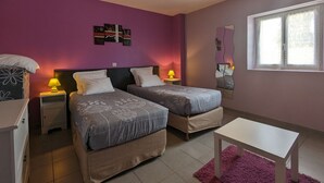 Un dormitorio con dos camas, una mesita de noche con lámpara, un aparador, una pequeña mesa y una ventana con persianas.