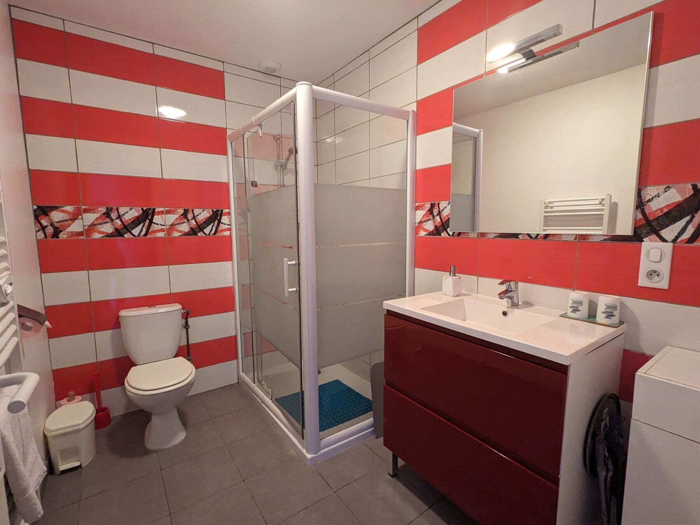 Ein Badezimmer mit einer gläsernen Duschkabine, einem weißen Waschbecken und einer roten Kommode.