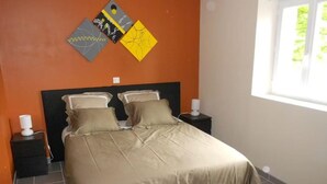 Una cama bien hecha en una habitación con pared naranja, una ventana y una mesita.