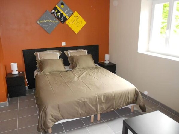 A neatly made bed in a room with an orange wall, a window, and a small table. - Cozy Room "Etoile Orange" - Ensuite Bathroom, Breakfast Included, Shared Kitchen and Lounge Area (Neuvy-Saint-Sépulchre)