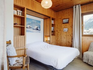 A wooden room with a bed, a chair, a shelf with books, a framed picture, and a window with a mountain view.