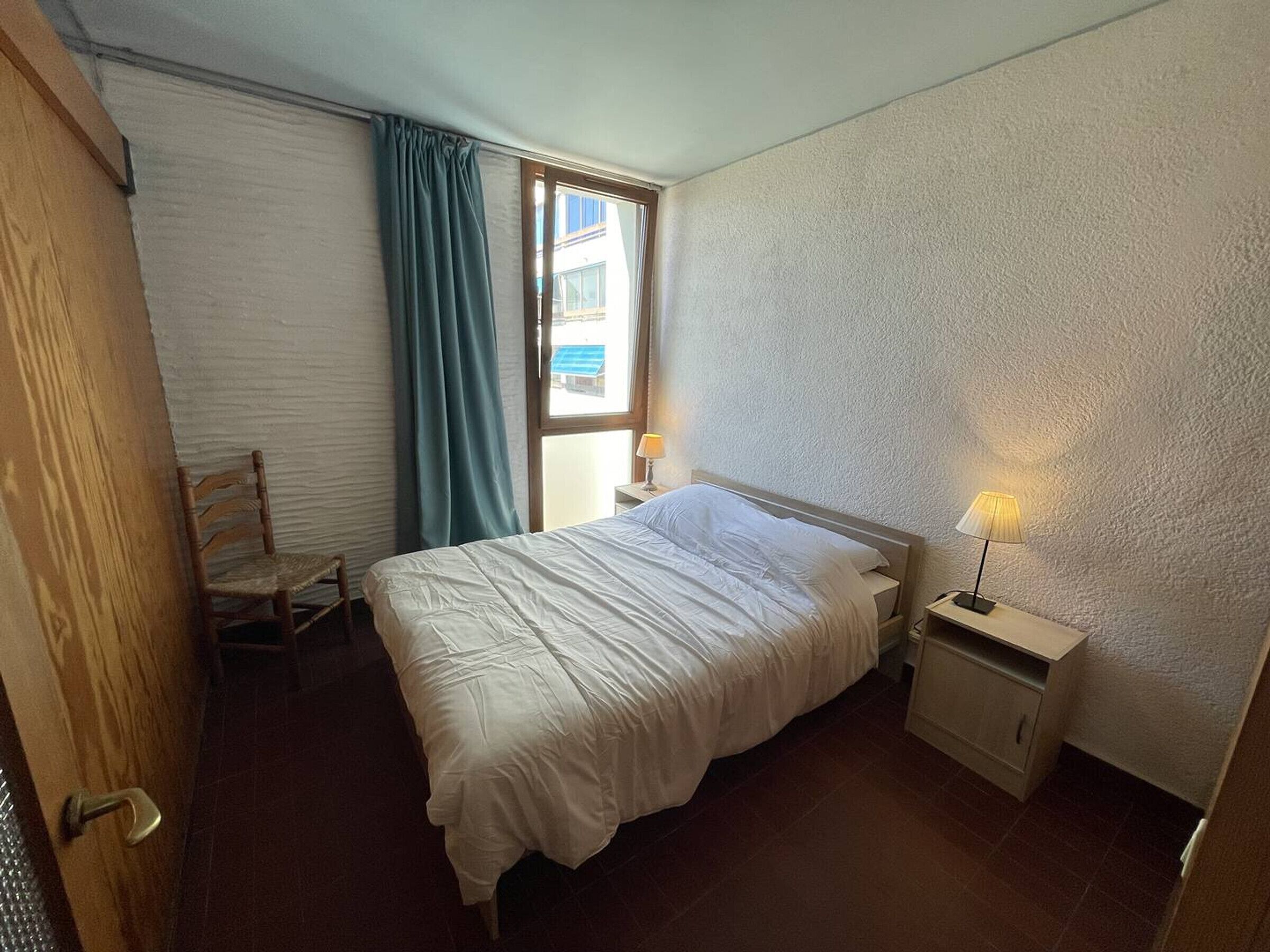 Een slaapkamer met een bed, een stoel, een nachtkastje met een lamp, een raam met gordijnen en een deur.