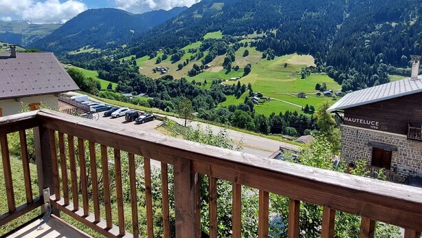 Vista de la montaña desde un balcón con baranda de madera, un edificio con la leyenda 'HAUTELUCE 1150' y un estacionamiento.