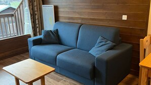 Una habitación acogedora con un sofá azul, una mesa de madera y vistas a una terraza de madera y colinas al exterior.