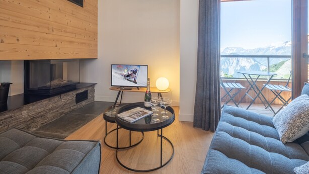 A modern living room with a fireplace, a television displaying a sports event, a small round table with a lamp, and a balcony with a mountain view. - New duplex, near slopes, 8 pax, parking (Huez)