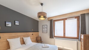 A bedroom with a wooden headboard, a bed with white linens, a wooden nightstand, a chair, a window with curtains, and a wall-mounted light fixture. - New duplex, near slopes, 8 pax, parking (Huez)