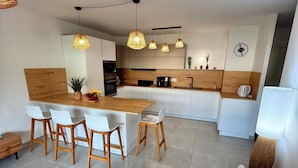 Une cuisine moderne avec des armoires blanches, un plan de travail en bois et des luminaires suspendus. On y trouve également un coin repas avec une table et des chaises en bois.
