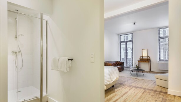 A modern room with a glass shower, a bed with a white duvet, a wooden floor, and a window with blinds. - Junior Suite in The Heart of Antwerp (Antwerpen)