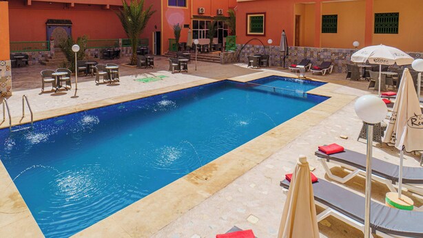 A swimming pool with lounge chairs and umbrellas, surrounded by a building with balconies and outdoor seating. - HOTEL LA VALLEE DES KASBAHS CHEZ ILYAS (Tinghir)