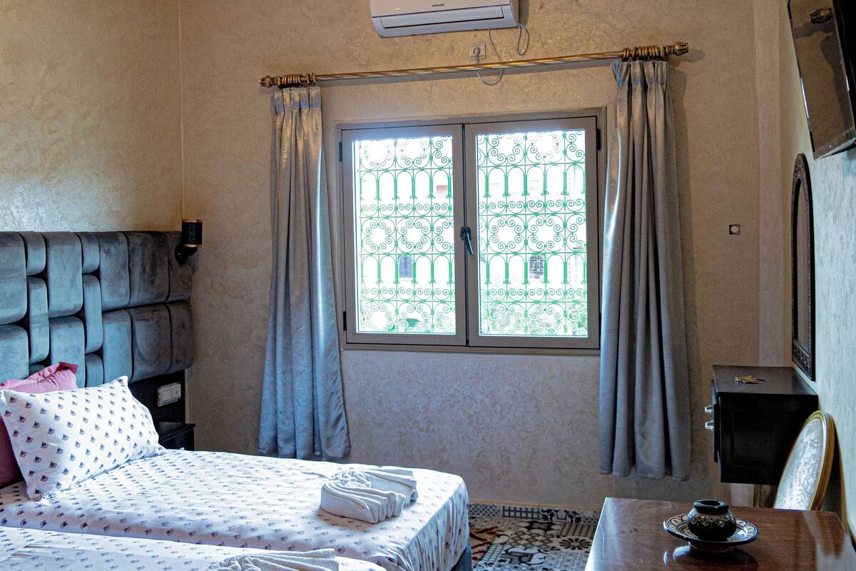 A hotel room with two beds, a desk, a chair, an air conditioning unit, a TV, and a window with curtains.